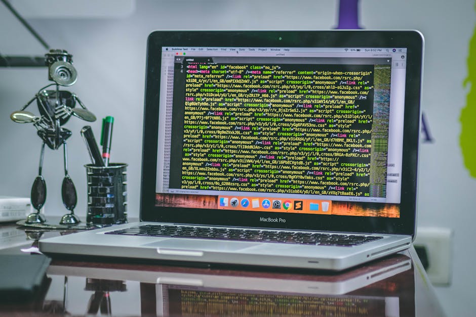 A close-up of a MacBook Pro laptop placed on a wooden desk, displaying a screen filled with lines of HTML code in various colors including green, yellow, and white. To the left of the laptop, there is
